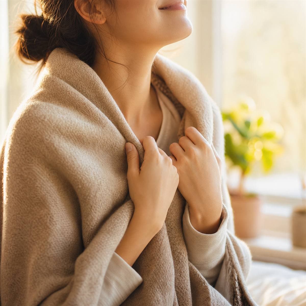 An adult gently wrapping a warm blanket around their own shoulders by a window, representing self-compassion and reparenting in trauma recovery